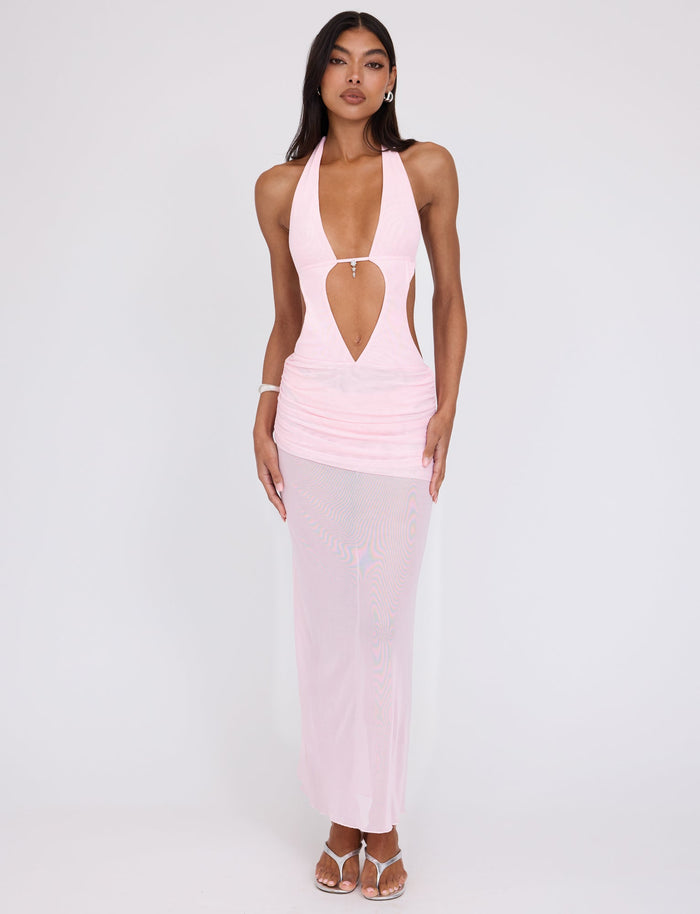 A woman stands against a plain background, wearing the TIGERMIST ESHA MAXI DRESS in Baby Pink, which features a light pink halter-top design with a keyhole cutout at the chest and a fitted, sheer skirt. She has long, dark hair and accessorizes with hoop earrings, a bracelet, and strappy heels.