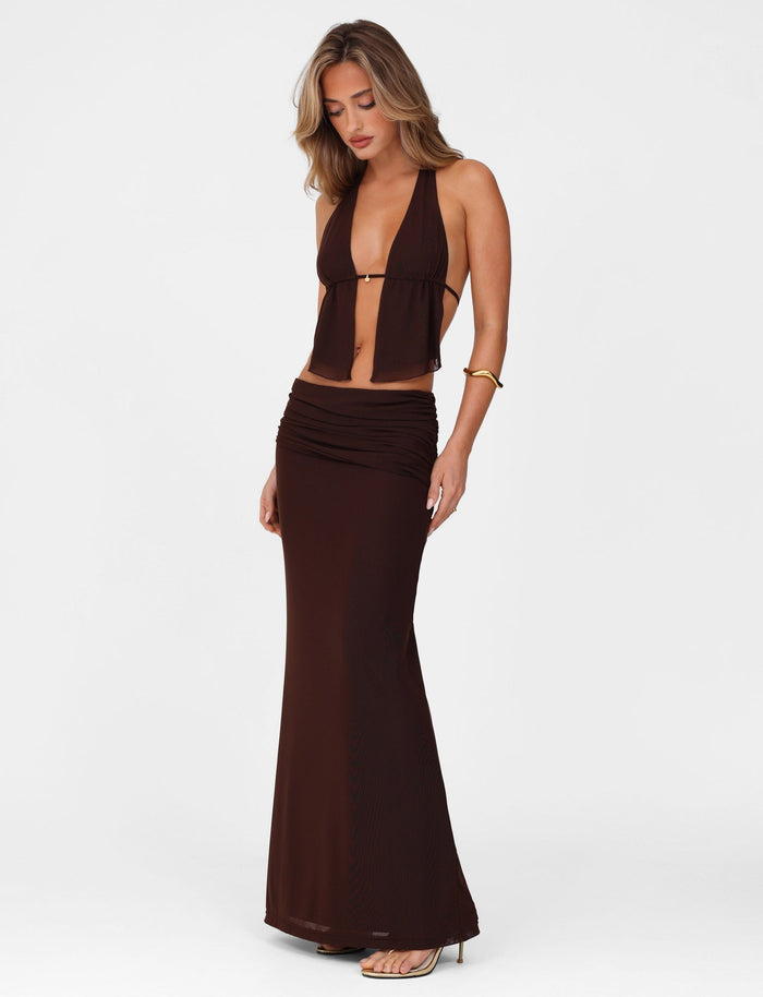 Against a white backdrop, a woman in the TIGERMIST chocolate ESHA MAXI SKIRT and dark brown halter top gazes downward. Her loose, wavy hair flows elegantly as she completes her look with high heels, an asymmetrical waistband, and a gold bracelet.