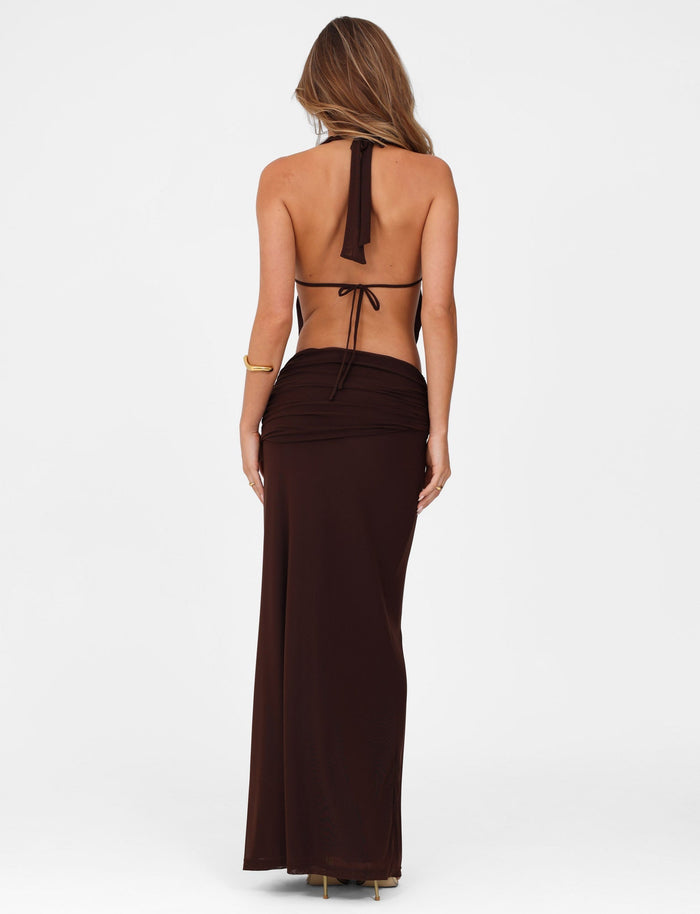 A woman with long hair, back to the camera, wears a chocolate Esha Maxi Skirt by Tigermist—a stunning backless outfit featuring a halter neck flowing elegantly to her feet. A gold bracelet adorns her wrist against the plain white background.