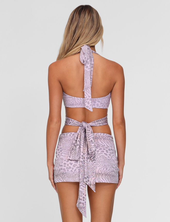 A woman with long blonde hair is seen from behind wearing the TIGERMIST EVANGELINE SKIRT - PINK : TIGER set, featuring a pink tiger print halter crop and matching bodycon mini skirt with large ties, against a plain white background.