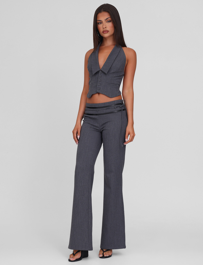 A woman with long dark hair wears the TIGERMIST FEDRA PANT - GREY, a pair of mid-rise tailored suit pants with side zippers, styled with a sleeveless deep V-neck gray vest and black high-heeled sandals against a light gray background.