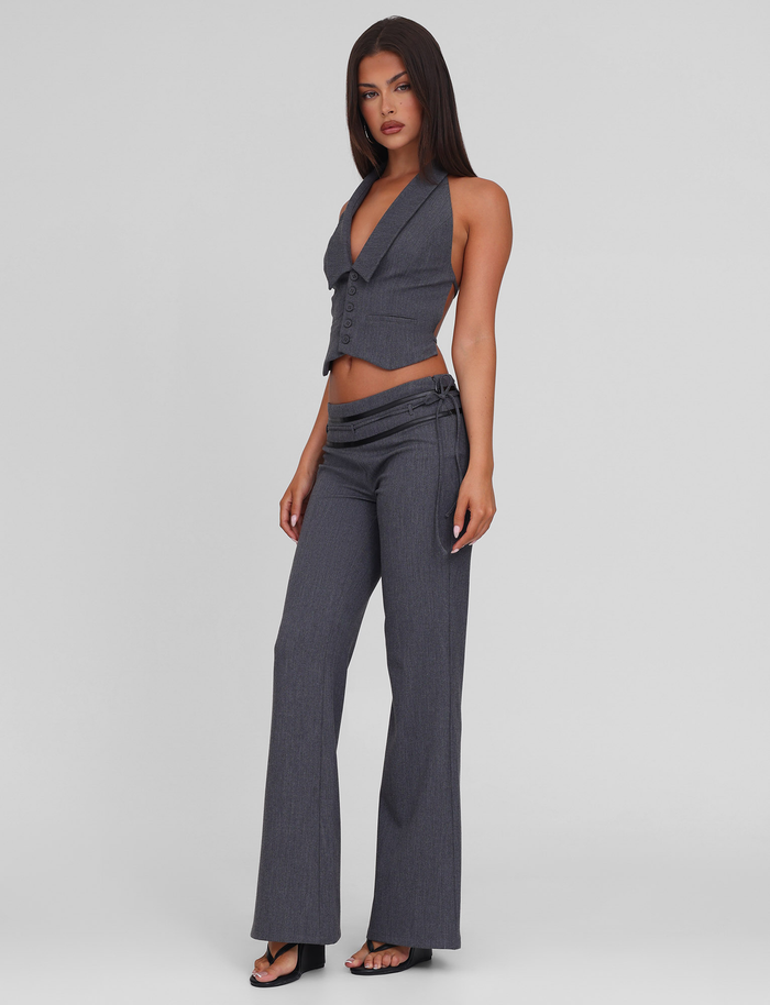 A woman stands against a plain background wearing TIGERMIST's FEDRA PANT in grey, styled high-waisted and tailored, paired with a matching fitted, sleeveless grey vest with PU leather trim and black heeled sandals.