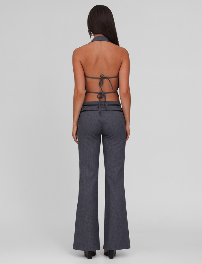 A woman with long dark hair faces away, wearing the TIGERMIST FEDRA PANT - GREY with a matching grey halter-style backless top tied with thin straps, set against a plain light grey background.
