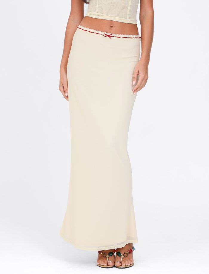A person is dressed in the Pale Yellow Fennel Skirt by Tigermist, a sheer piece with lace trim details at the waist. They pair it with a cropped sleeveless top and sandals adorned with decorative straps. The plain white background highlights the outfit's elegance.
