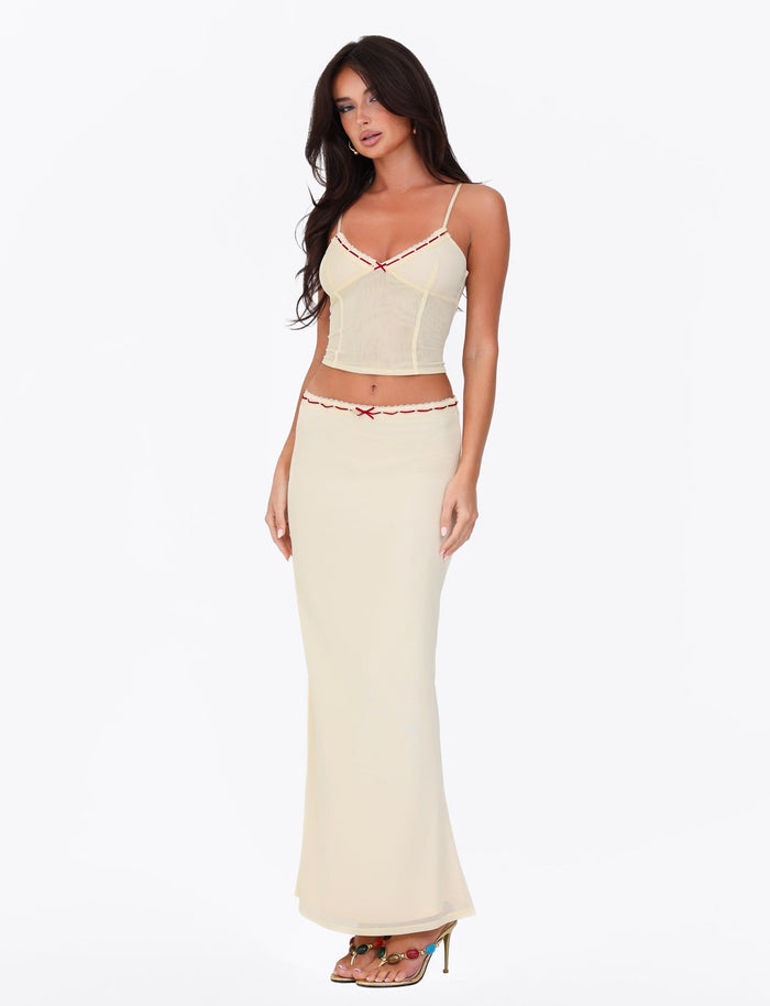 Dressed in the FENNEL SKIRT by TIGERMIST, a person exudes elegance against a white background. The pale yellow hue of the skirt is complemented by its form-flattering fit, while delicate lace details highlight its charm. Paired effortlessly with high-heeled sandals, the ensemble elegantly enhances their silhouette.