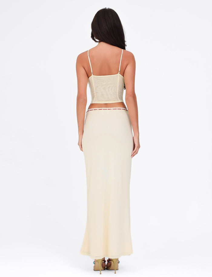 A woman with long dark hair is wearing a beige, form-fitting, sleeveless top paired with the FENNEL SKIRT in pale yellow by TIGERMIST. The high stretch fabric accentuates her silhouette as she stands with her back to the camera against a plain white background.