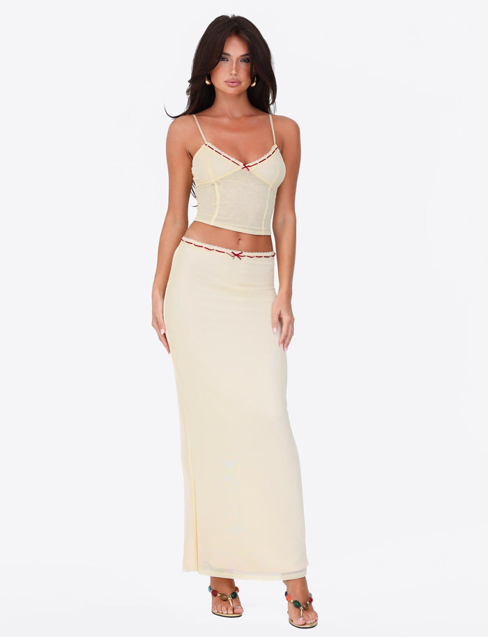 A woman models against a white backdrop, showcasing the TIGERMIST FENNEL SKIRT in pale yellow, complemented by delicate lace trimming, red accents, and a coordinating light yellow strappy top. Her long dark hair cascades down as she completes the look with stylish sandals.