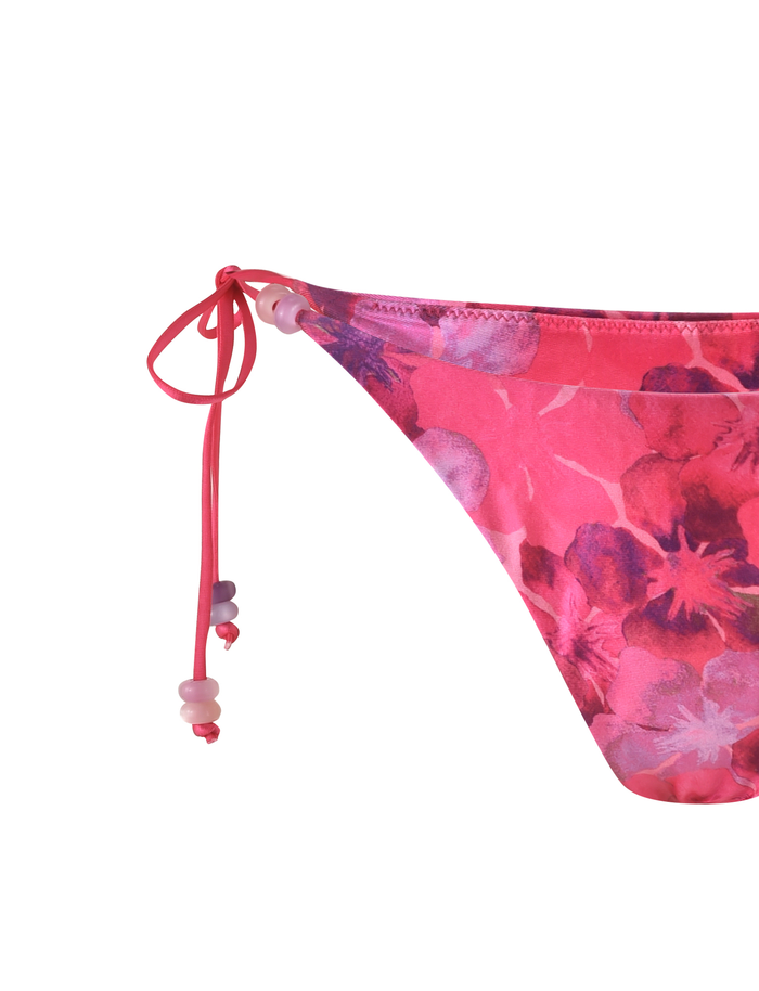Close-up of the TIGERMIST FINLEA BIKINI BOTTOM - PINK : HIBISCUS, featuring a pink floral cheeky cut with side tie strings and decorative beads, crafted from soft elastane fabric against a white background.