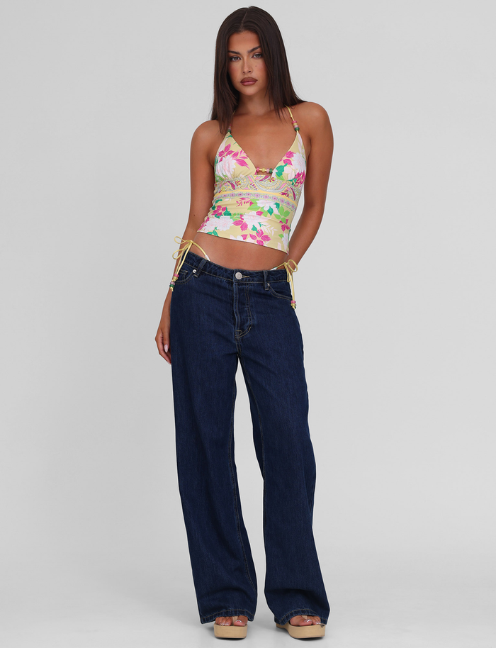 A woman with straight brown hair wears the TIGERMIST FINLEA TANKINI TOP - PAISLEY BORDER, paired with wide-leg dark blue jeans and platform sandals, standing against a plain light gray background.