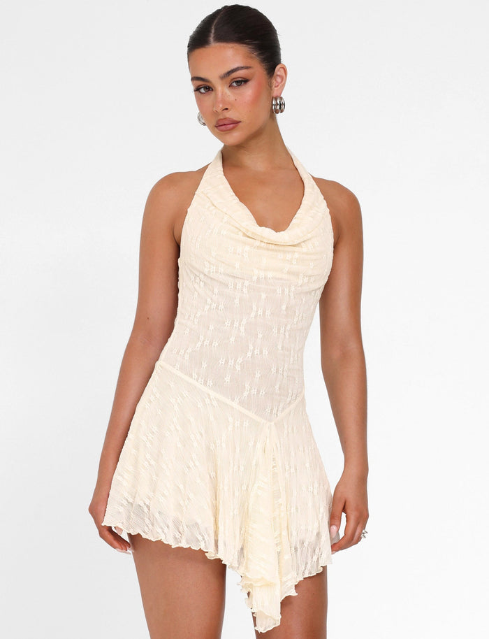 A woman wears the TIGERMIST FIORELLA MINI DRESS in cream lace with a halter neck and asymmetrical hem, standing against a white background and looking at the camera.
