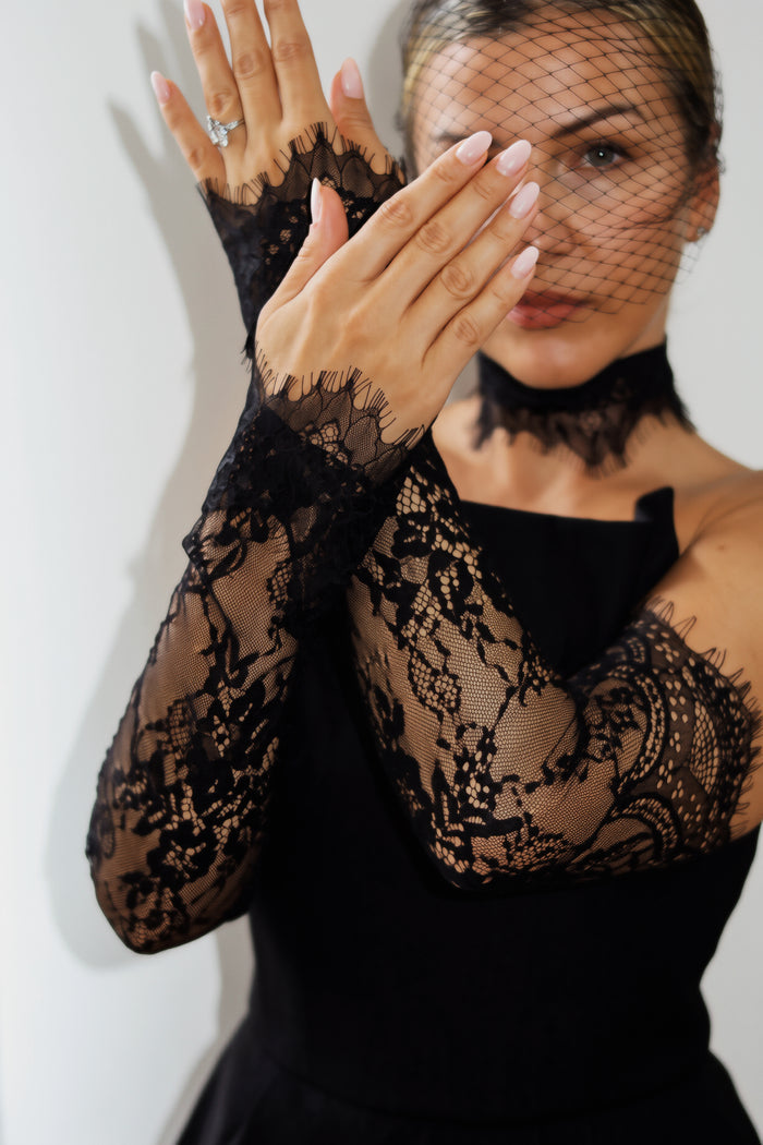 A woman wearing long black lace gloves with a matching black fishnet veil and black dress against a plain light background.