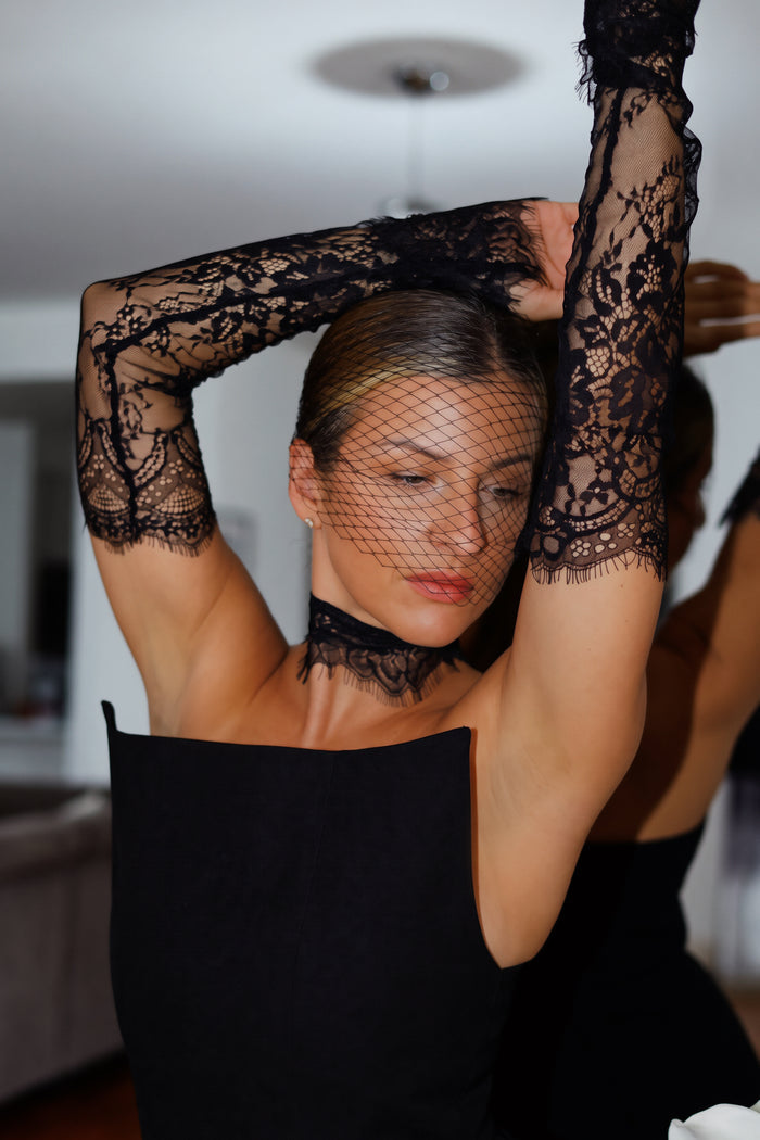 A woman wearing a black lace long-sleeve top with a matching black lace choker and fishnet head covering, posing with her arms raised in an indoor setting.