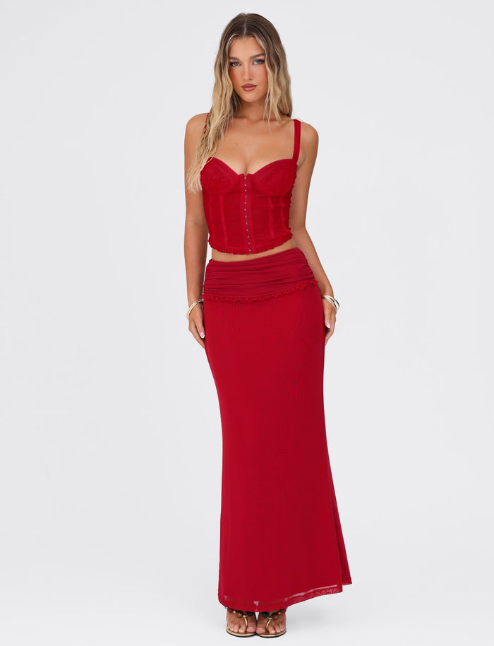 A woman with long, wavy hair is wearing a red, sleeveless corset top and the TIGERMIST Ginny Maxi Skirt in Cherry Red. She has minimal accessories and is standing against a plain, light-colored background.