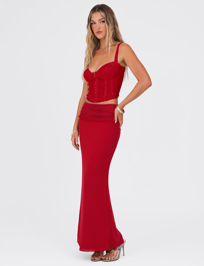 A woman stands against a plain white background, wearing a fitted, vibrant red corset top and the GINNY MAXI SKIRT - RED : CHERRY RED from TIGERMIST—a matching red maxi length skirt with a slight flare at the bottom. She has long, wavy blonde hair and is posing with one hand on her hip and the other trailing at her side.