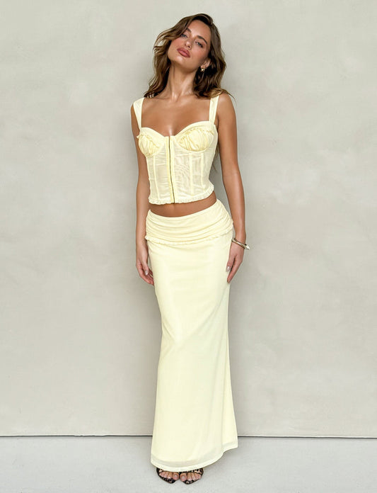 A woman is posing against a plain backdrop, wearing a fitted yellow corset top and the TIGERMIST GINNY MAXI SKIRT - LEMON, which features a wide waistband. She stands with her head tilted and her hair flowing freely.