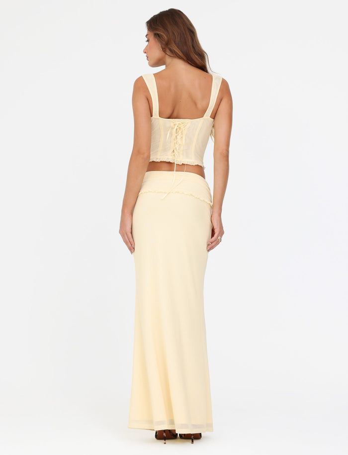 A woman stands with her back to the camera, wearing a cream-colored, sleeveless top featuring lace-up details paired with the TIGERMIST GINNY MAXI SKIRT in yellow lemon. Her long brown hair cascades down against the plain white background.