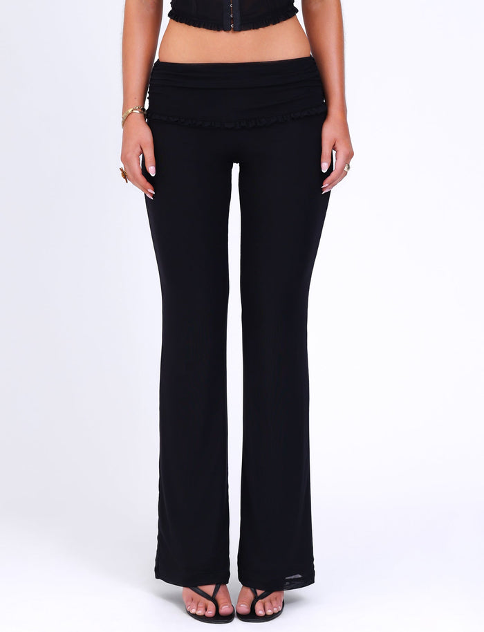 A person is adorned in TIGERMIST's GINNY PANT - BLACK, complemented by black open-toe sandals, standing against a white backdrop. The individual, also wearing a cropped black top, is photographed from the waist down to emphasize the chic flared silhouette.