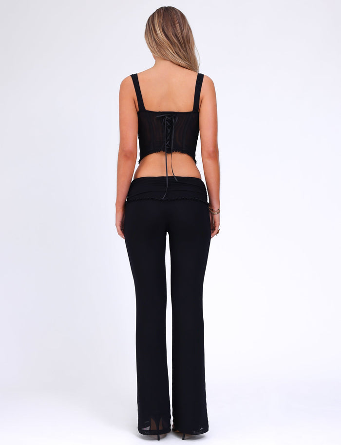 A person with long blonde hair stands facing away from the camera against a plain background. They are wearing a black, sleeveless top with a lace-up back and the GINNY PANT - BLACK from TIGERMIST, which features a low-rise waist and flared silhouette.