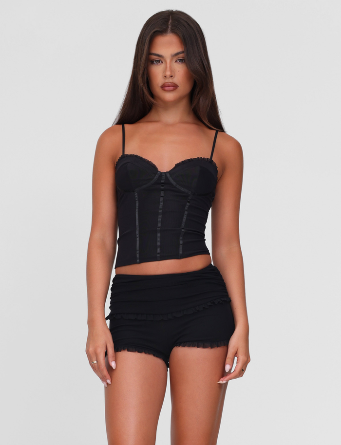 A woman with long dark hair stands against a light gray background, wearing TIGERMIST's GINNY SHORT - BLACK and a fitted black camisole, showcasing the chic bodycon silhouette as she looks directly at the camera with a neutral expression.