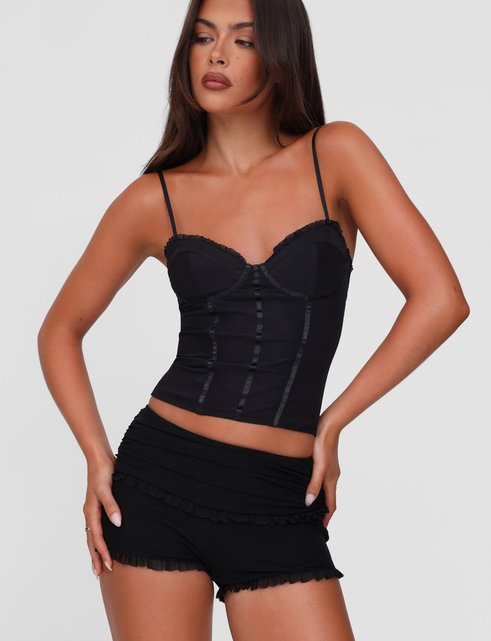 A woman with long brown hair models the TIGERMIST GINNY SHORT - BLACK, styled with a fitted black camisole, posing with one hand on her hip against a plain light gray background.