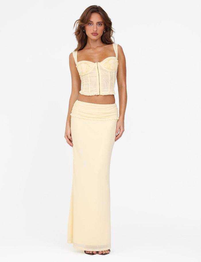 A woman stands against a plain white background wearing the TIGERMIST GINNY TOP in Lemon, featuring a sweetheart neckline, paired with a matching long skirt. Her long, wavy hair flows gently around her face as she gazes softly with her hands resting by her sides.