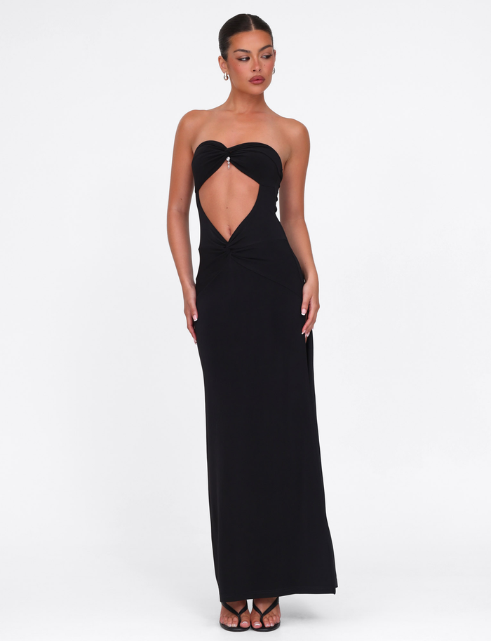 A woman poses against a white background wearing the TIGERMIST GRAZIA DRESS in black, a stretch bandeau maxi dress with a central midriff cutout. Her hair is pulled back and she pairs the look with black sandals.