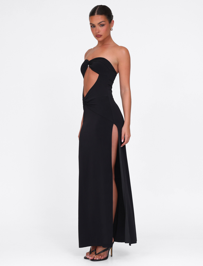 A woman stands against a plain white background wearing the TIGERMIST GRAZIA DRESS - BLACK, a strapless maxi dress featuring stretch fabric, front cut-out, high side slit, and paired with black high heels.