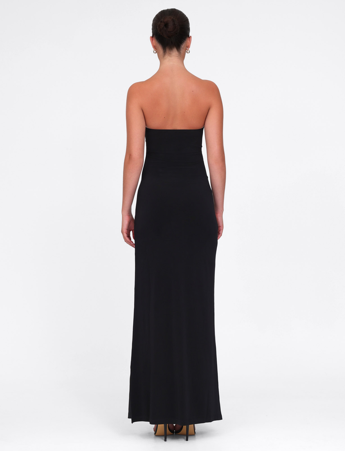 A woman stands with her back to the camera in the TIGERMIST GRAZIA DRESS - BLACK, a sleek stretch bandeau maxi dress, styled with high heels and a neat bun against a plain white background.