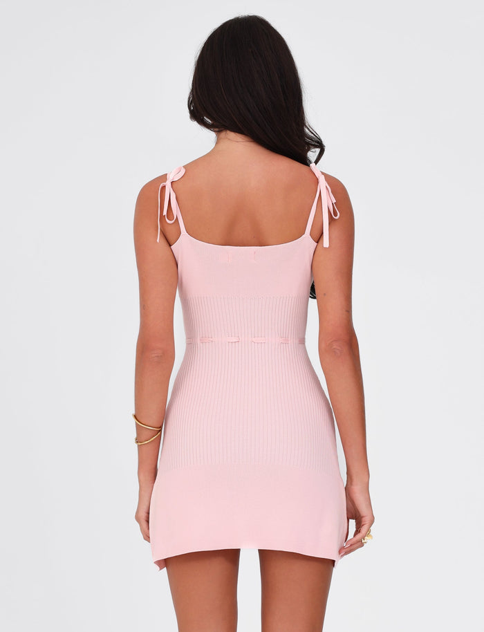 A woman with long brown hair is facing away, wearing the TIGERMIST GREER DRESS - PINK in baby pink. The dress showcases true-tie shoulder straps, and her relaxed arms are adorned with bracelets. The plain white background complements the knit fabrication of the dress.