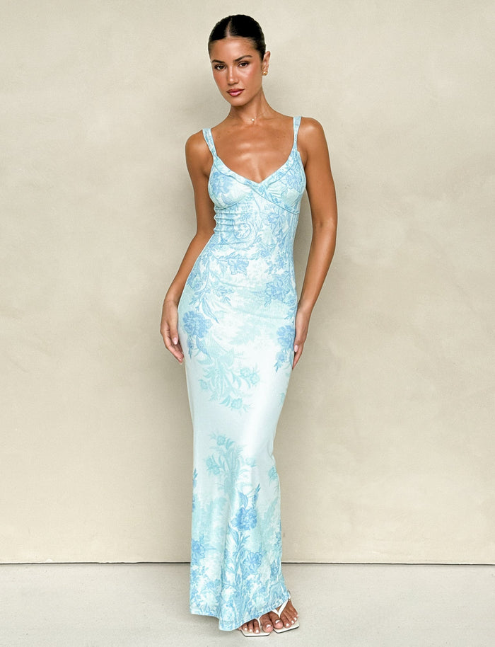 A woman poses in front of a neutral backdrop, wearing the TIGERMIST GUILIANNA FRILL MAXI DRESS - BLUE : FLORAL : TAPESTRY, a form-fitting and sleeveless piece crafted from high stretch fabric. Her hair is pulled back and she pairs the elegant V-neck design with white sandals, highlighting its adjustable frill shoulder straps that add an extra touch of charm.