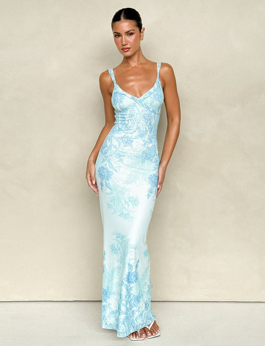 A woman poses in front of a neutral backdrop, wearing the TIGERMIST GUILIANNA FRILL MAXI DRESS - BLUE : FLORAL : TAPESTRY, a form-fitting and sleeveless piece crafted from high stretch fabric. Her hair is pulled back and she pairs the elegant V-neck design with white sandals, highlighting its adjustable frill shoulder straps that add an extra touch of charm.