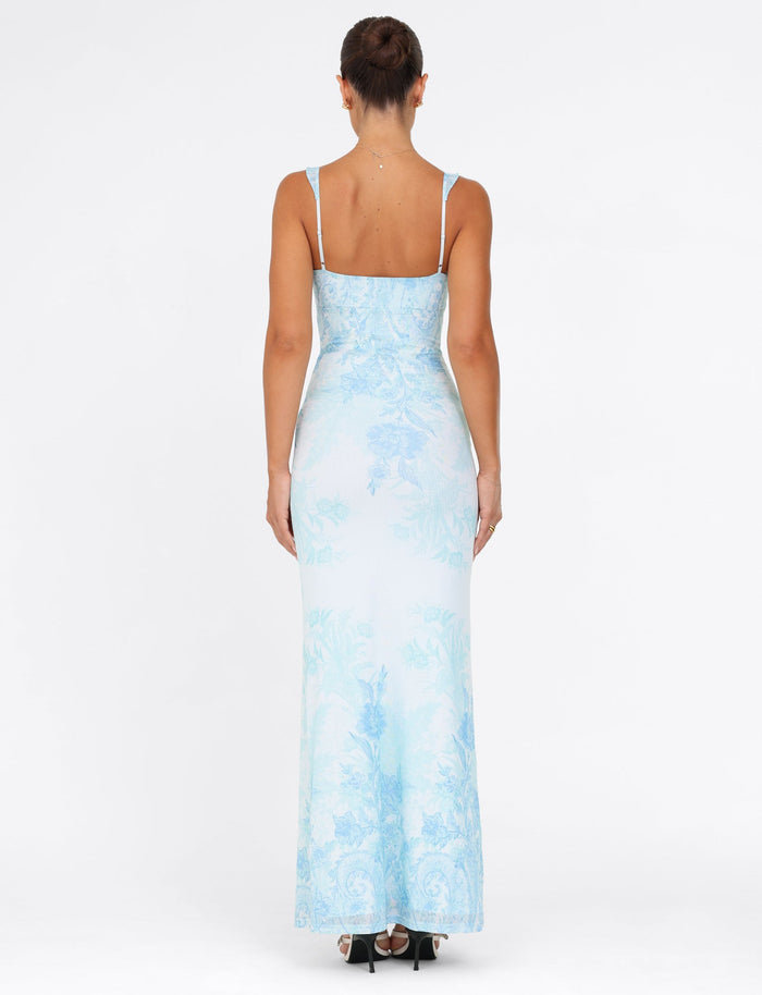 A person stands with their back to the camera, dressed in the TIGERMIST GUILIANNA FRILL MAXI DRESS - a long, blue floral tapestry dress featuring adjustable frill shoulder straps and a V-neckline. The fitted silhouette highlights their neat updo and strappy heels against a plain white background.