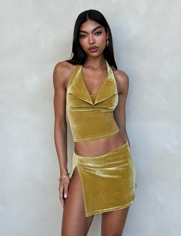A woman with long, dark hair poses against a neutral background, wearing the HARLOWE SKIRT - GOLD and a matching yellow velvet halter top by TIGERMIST. The sleeveless outfit emphasizes her toned midriff as she accessorizes with gold jewelry, including earrings and bracelets.