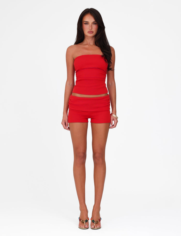 The woman, wearing the HARRIETT RIB TUBE TOP in red from TIGERMIST paired with matching shorts, stands elegantly against a plain white background. Her bodycon silhouette is accentuated by her heels and bracelet, with long dark hair cascading down her shoulders.