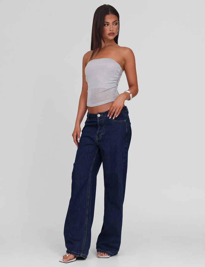 A woman models the TIGERMIST HARRIETT TUBE TOP - SILVER with dark blue wide-leg jeans, standing against a plain light gray background, gazing left with one hand by her side and the other touching her hip.