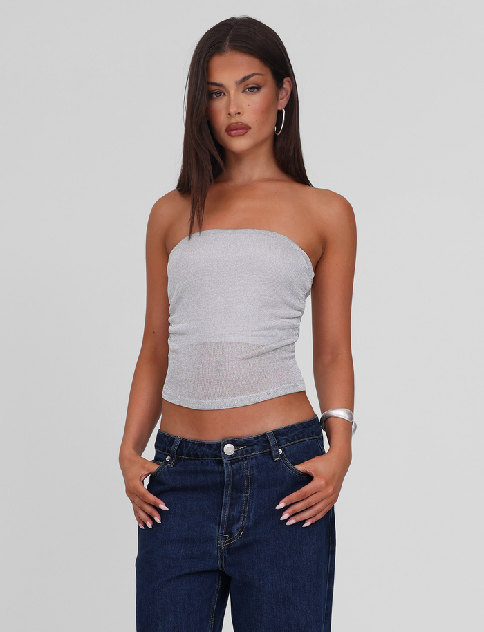 A woman with straight brown hair wears the TIGERMIST HARRIETT TUBE TOP in silver and dark blue jeans, standing with her hands in her pockets against a plain light gray background.