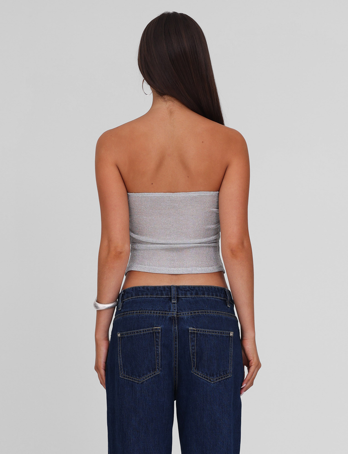 A woman with long brown hair stands facing away, wearing the TIGERMIST HARRIETT TUBE TOP in silver and blue jeans, set against a plain light gray background.