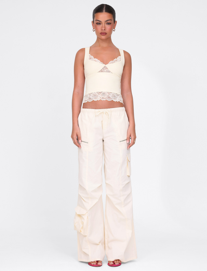 A woman stands against a white background wearing the TIGERMIST HERMIONE PANT in Soft Butter, paired with a cream lace-trimmed crop top and red open-toe heels. Her tied-back hair and neutral expression complete the relaxed vibe.