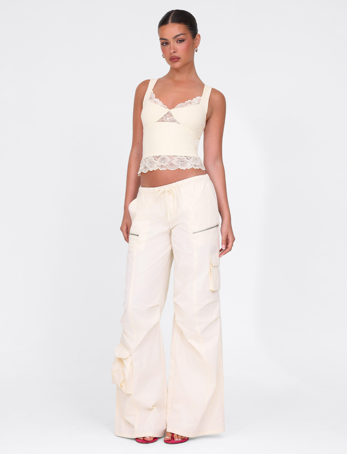 A woman poses against a white background in TIGERMIST's HERMIONE PANT - YELLOW : BUTTER : SOFT BUTTER, styled with a cream lace-trimmed crop top, tied-back hair, and red open-toe heels for a chic, casual vibe.