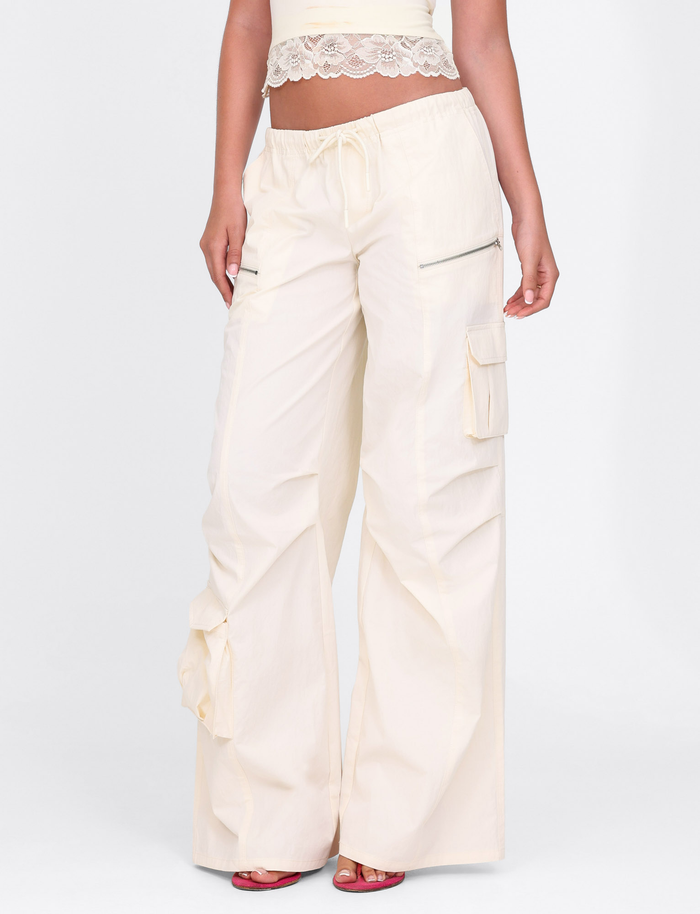 A person wears TIGERMIST's HERMIONE PANT in Soft Butter, featuring a drawstring waist and pockets, styled with a cropped lace-trim top and red heeled sandals, standing against a plain white background.