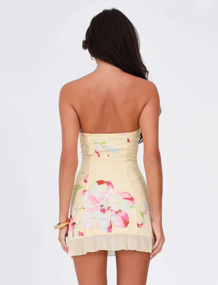The woman with long dark hair is wearing the HOLLIS DRESS - YELLOW : FLORAL : FEMME FLORAL by TIGERMIST, a strapless corset dress featuring pink and green floral patterns. She is standing against a plain white background, viewed from the back.