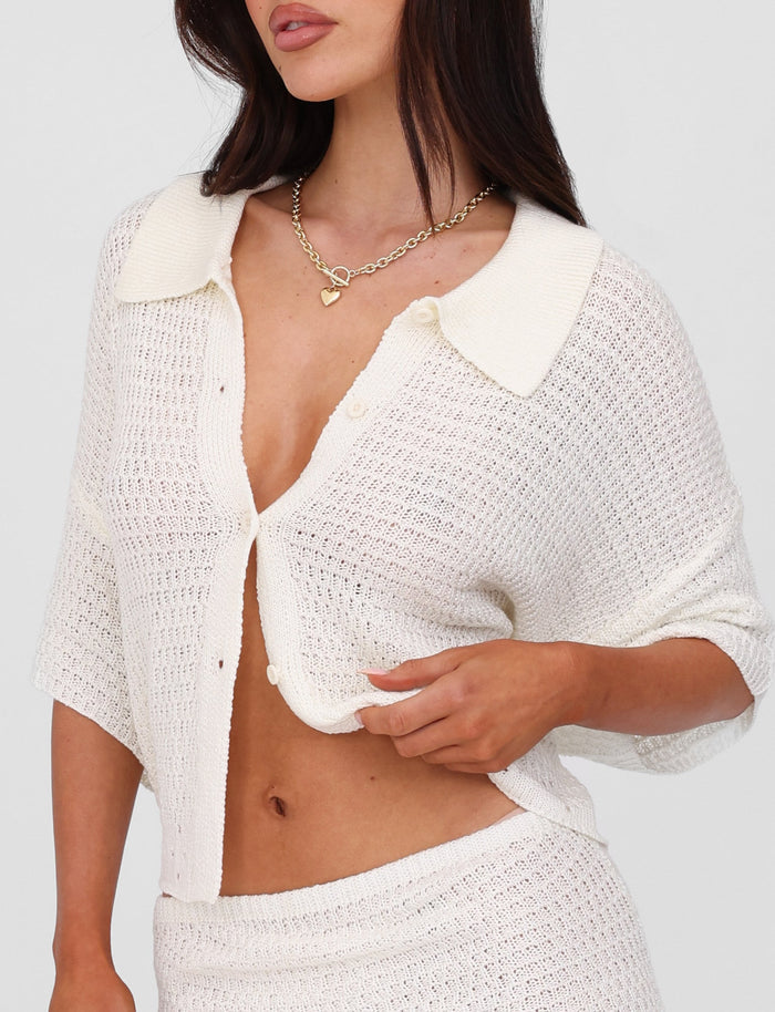 A woman with long dark hair wearing the oversized fit JANAI TOP - WHITE : CREAM from TIGERMIST, along with a heart-shaped necklace. The partially buttoned, collared neckline cardigan is held gently as she tugs at its hem, and it’s paired perfectly with a matching knit skirt.
