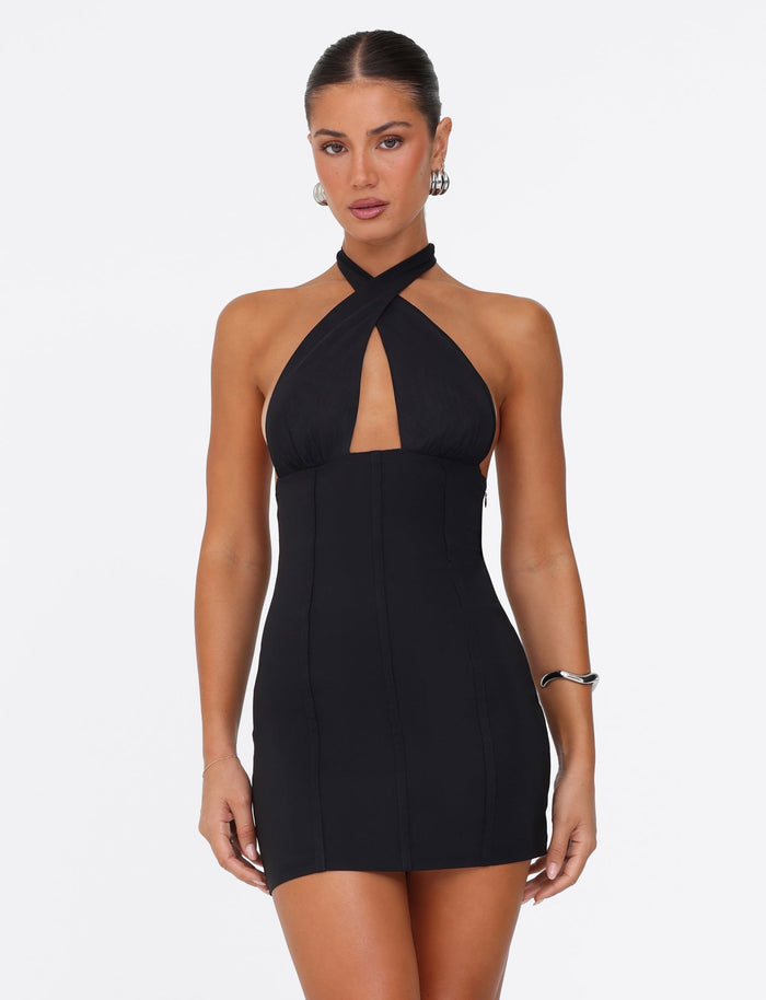 A woman poses against a white background wearing the JANECE MINI DRESS in black by TIGERMIST, which features an elegant mesh-lined bust and a front cutout. Her hair is pulled back, accentuating her hoop earrings and the silver cuff bracelet on her right arm.