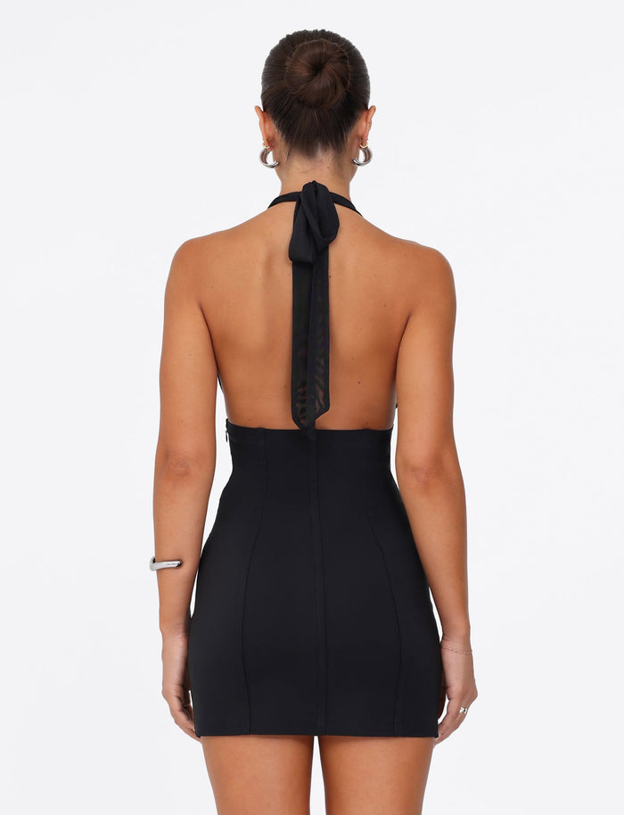 A woman stands with her back to the camera, wearing the JANECE MINI DRESS - BLACK by TIGERMIST. Her hair is styled in a bun, and she accessorizes with hoop earrings. She holds one arm by her side while the other is bent, all set against a plain white background that accentuates the dress's elegance and form-fitting design.