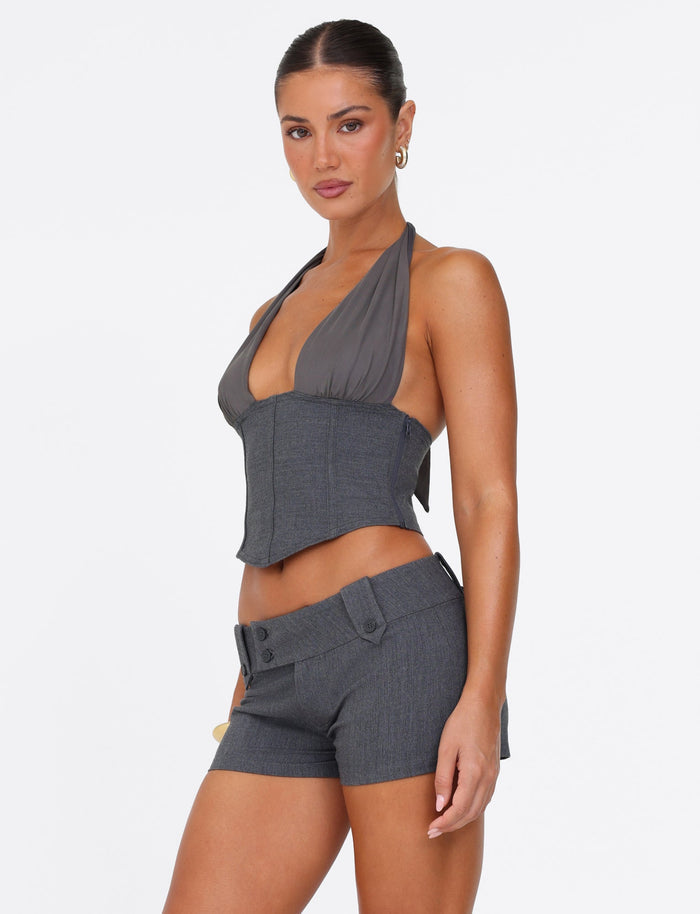 A person modeling a stylish outfit showcases the dark gray Janece Top by Tigermist, featuring a plunging neckline, paired with grey suiting shorts adorned with a chic belt detail. The individual poses confidently against a plain white background.