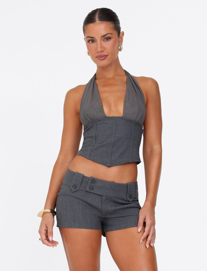 A woman stands confidently against a plain white background, wearing the JANECE TOP - GREY from TIGERMIST, crafted with grey suiting fabrication and paired perfectly with matching shorts. Her sleek hairstyle, hoop earrings, and bracelet on her left wrist enhance her poised and assured expression.