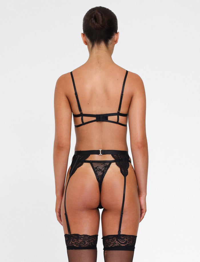 A person faces away from the camera, highlighting the JORDAN SET - BLACK by TIGERMIST, featuring a lace underwire bra, thong, garter belt, and stockings. Their hair is styled in a bun against a plain white background.