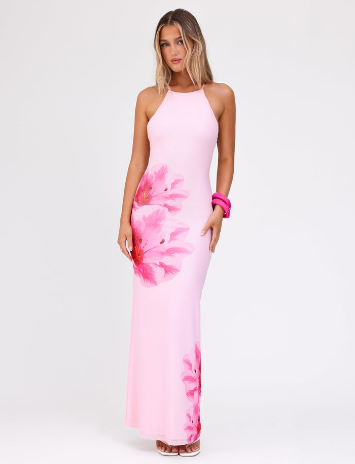 A woman wearing the TIGERMIST KARTER DRESS in baby pink floral print, a sleeveless maxi dress with large prints, stands against a plain white background. She has long, wavy blonde hair and is accessorized with a pink bangle on her right wrist. Completing her look are pink high-heeled shoes.