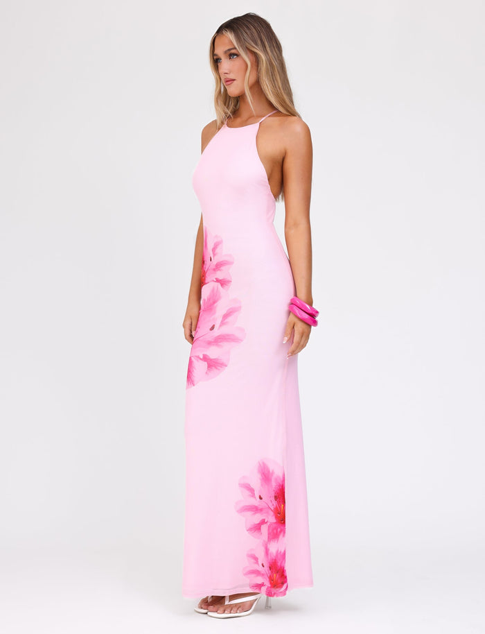 A woman stands against a plain white background, wearing the TIGERMIST KARTER DRESS in baby pink with a maxi length and floral mesh fabrication. She has long, wavy hair, accessorized with pink bangles on her wrist and white high-heeled sandals.
