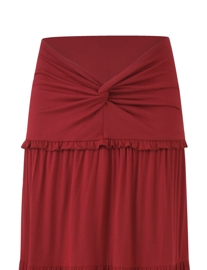 The KATYA MAXI SKIRT by TIGERMIST is a red maxi skirt made from high stretch fabric, featuring a wide waistband with a twisted knot detail and a small ruffled trim.